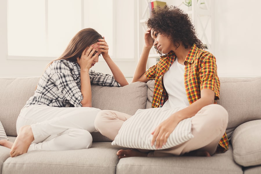 Two distraught female friends sitting on a couch discussing how to stop enabling behavior.