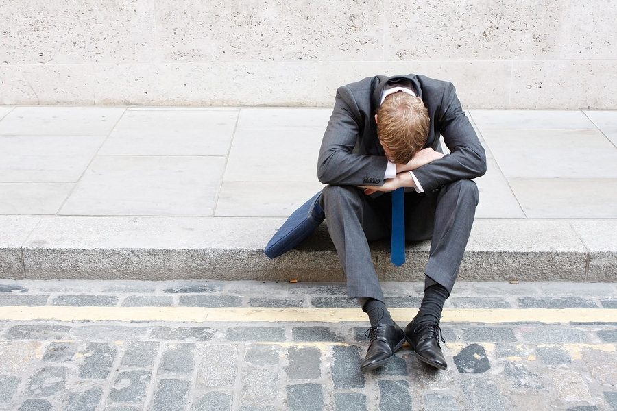 A businessman sits on the curb with his head and arms resting on his knees.