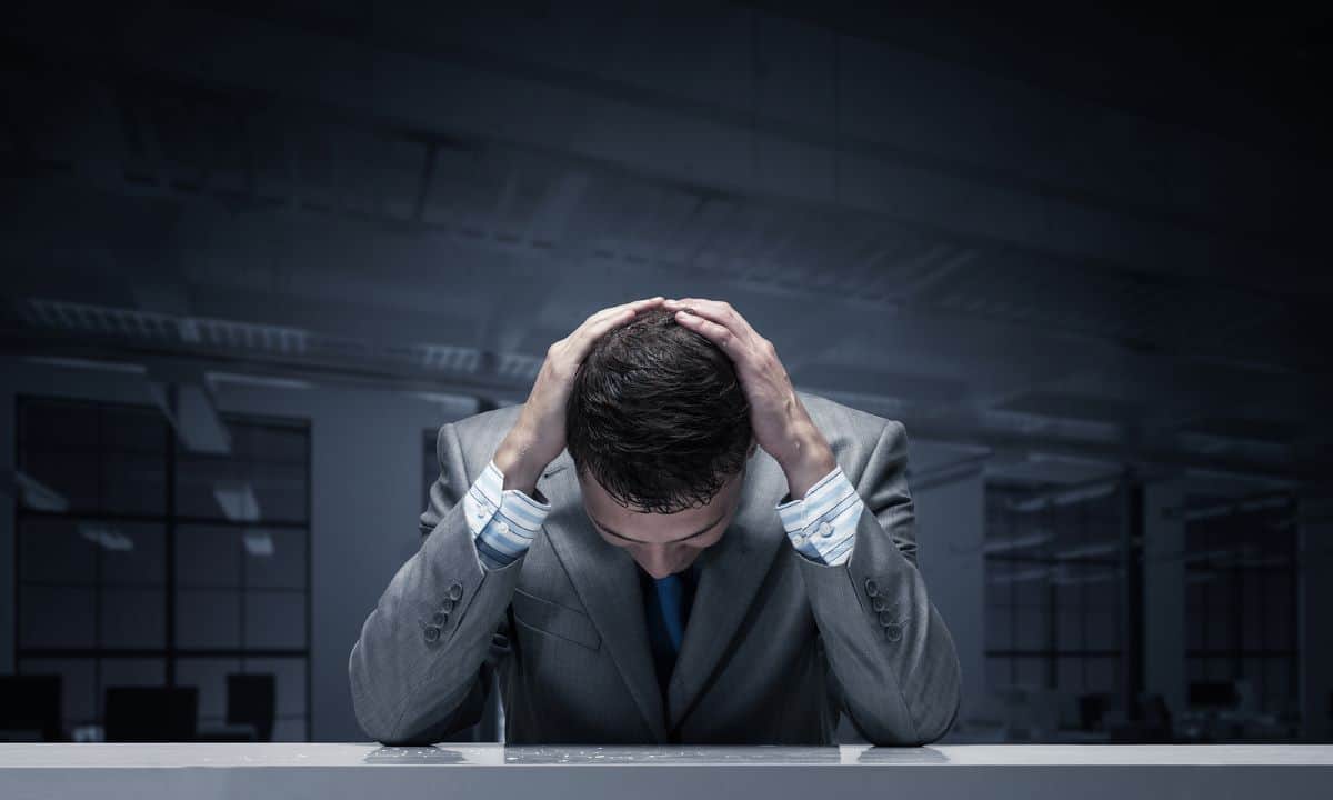 A stressed businessman sits at his desk with his hands on his head.