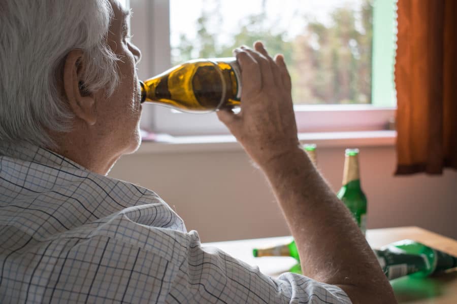 An older man sits at a table by a window binge drinking beer