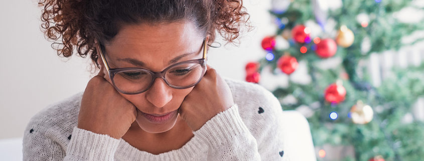 A woman sitting by a Christmas tree, struggling with holiday stress.