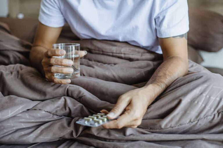 A man’s hand holding a glass of water in one and a packet of pills in another, wondering how Adderall abuse is treated.