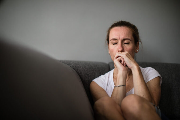 A depressed, mid-aged woman sits on her couch with her eyes closed and hands clasped close to her mouth.