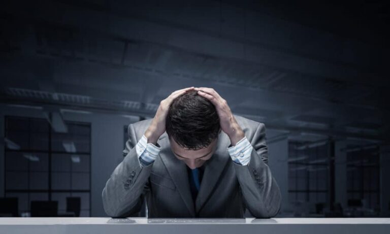A frustrated businessman sits at his desk with his hands on his head thinking how addictive heroin is.